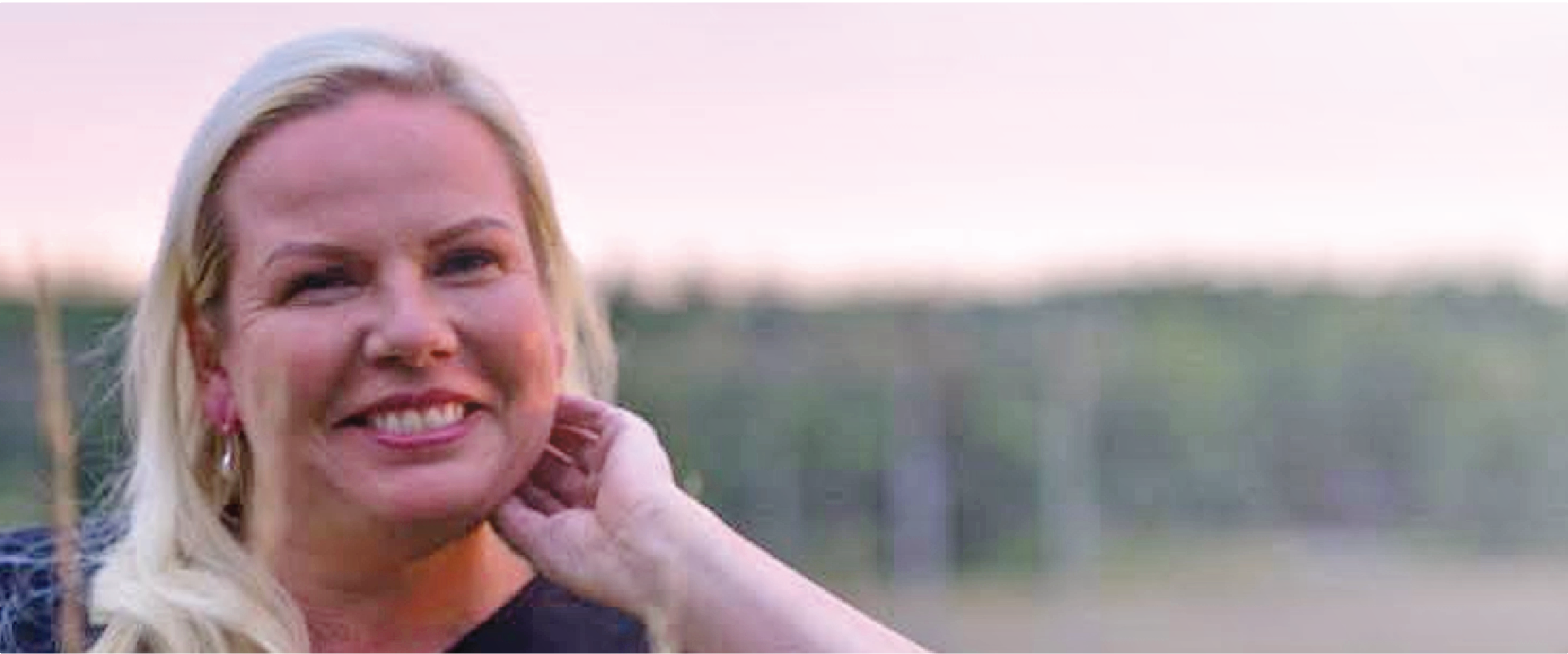 A blonde woman smiles warmly with her hand to her neck, soft sunset tones behind her — a re:you moment of calm, content connection.
