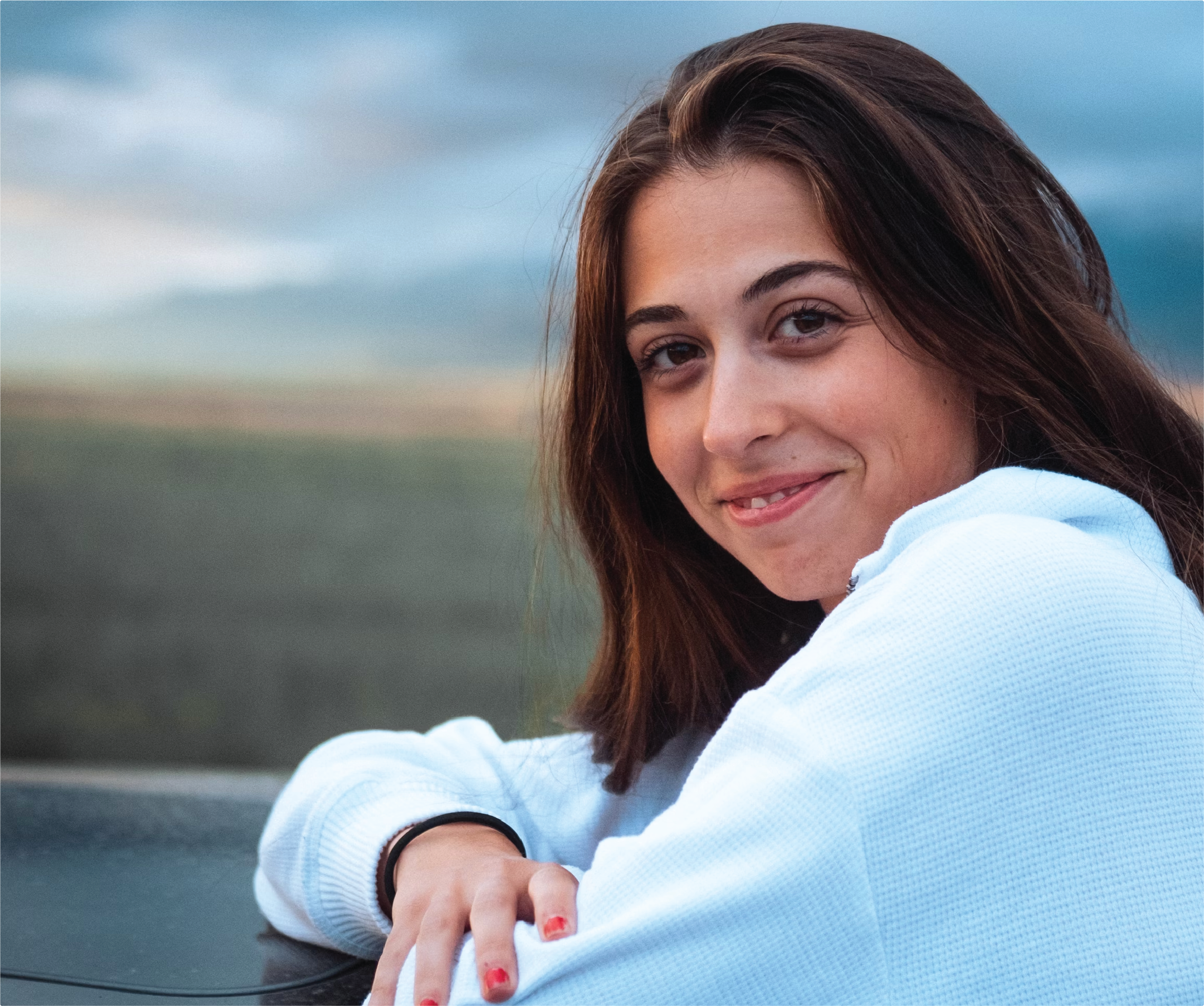 A young woman in a light blue sweater smiles softly at the camera during golden hour — a re:you moment of grounded warmth and quiet confidence.
