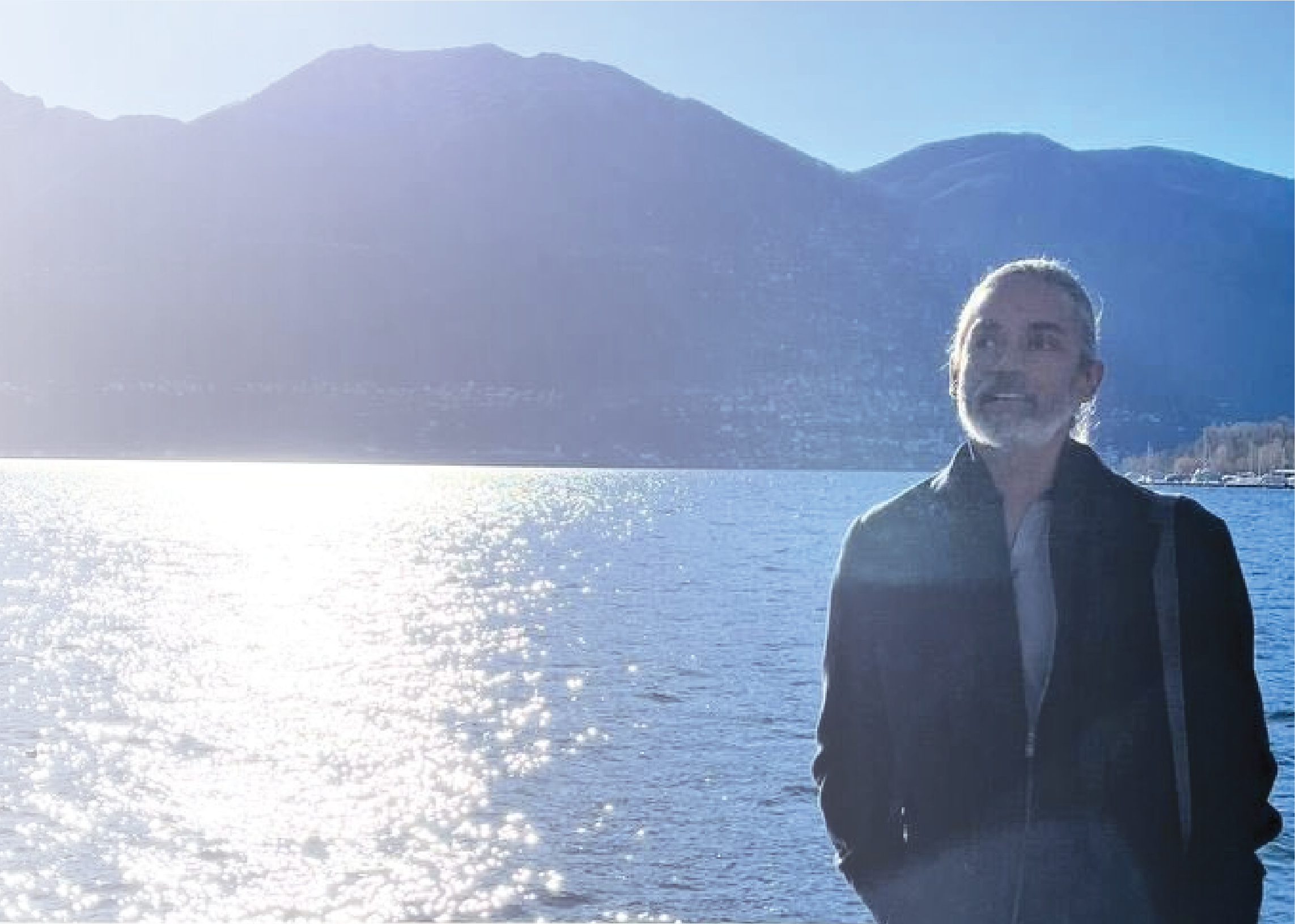 A silver-haired man stands by a sparkling lake at sunset, mountains behind him, fully in his own energy — a re:you moment of peace and presence.